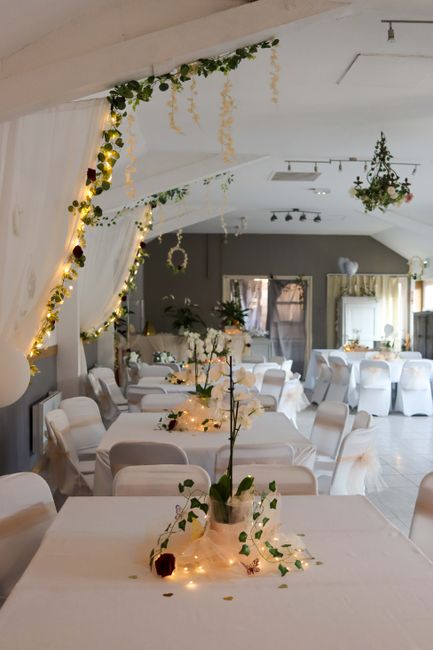 An elegantly decorated indoor venue featuring long banquet tables draped with white tablecloths, adorned with small floral arrangements and fairy lights. The floral arrangements include white roses, l