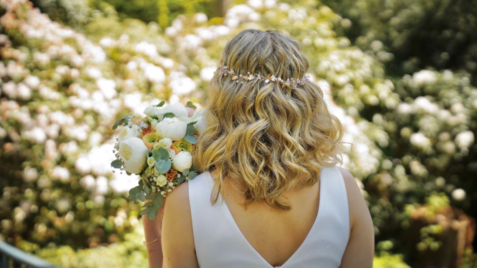 A woman with shoulder-length, wavy blonde hair adorned with a delicate floral headband is holding a bouquet of fresh wedding flowers close to her shoulder. The bouquet features white peonies, soft pin