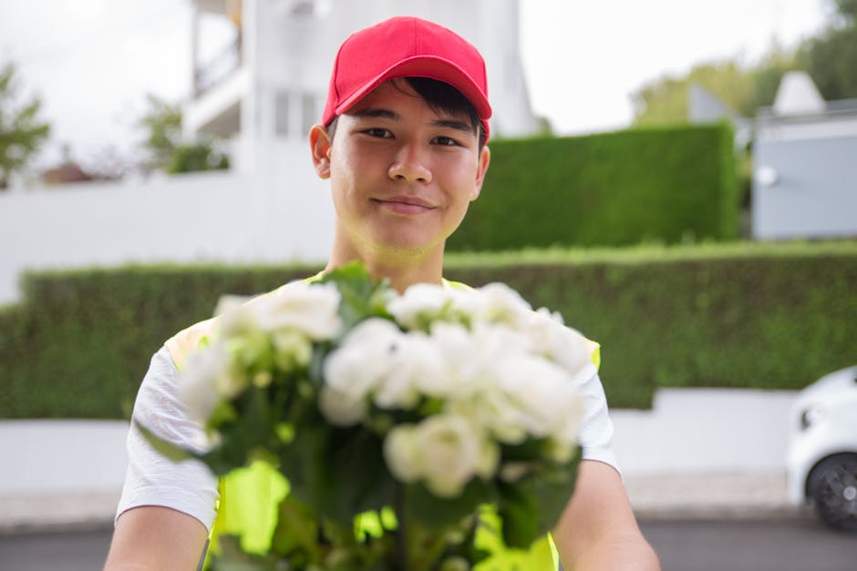 A young male flower delivery courier wearing a red cap and a high-visibility vest is smiling and holding out a bouquet of fresh white roses and green foliage towards the camera. The bouquet is wrapped
