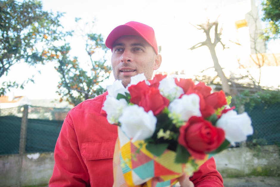 A male flower delivery person wearing a red cap and red jacket is holding a vibrant bouquet of fresh flowers, including red and white roses with lush green leaves, wrapped in colorful patterned paper.
