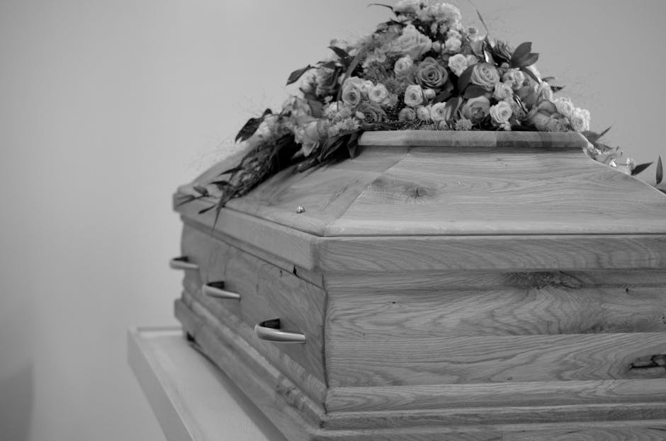 A wooden funeral casket with a polished, natural wood grain finish, adorned with a large floral arrangement of roses, lilies, and other mixed flowers in soft pastel shades. The casket is placed on a w