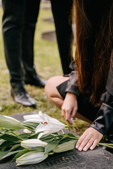 A person kneeling on the ground, adjusting a bouquet of white lilies with long green stems and leaves, which is placed on a flat surface in a memorial setting. The lilies are fresh, with smooth petals