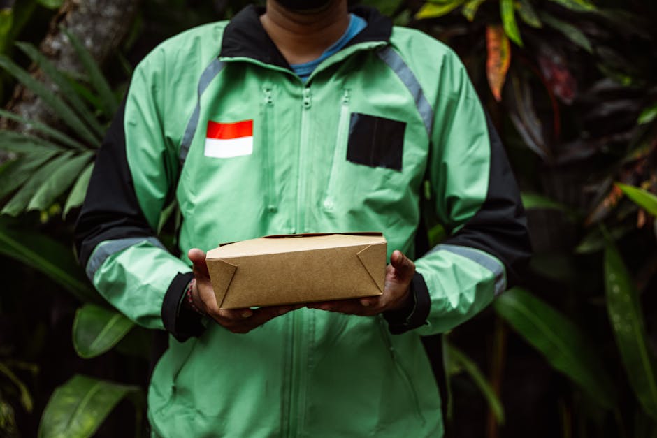 A person wearing a green jacket with black and grey accents, featuring a small Indonesian flag patch and a black rectangular patch, is holding a rectangular brown paper package with folded corners. Th